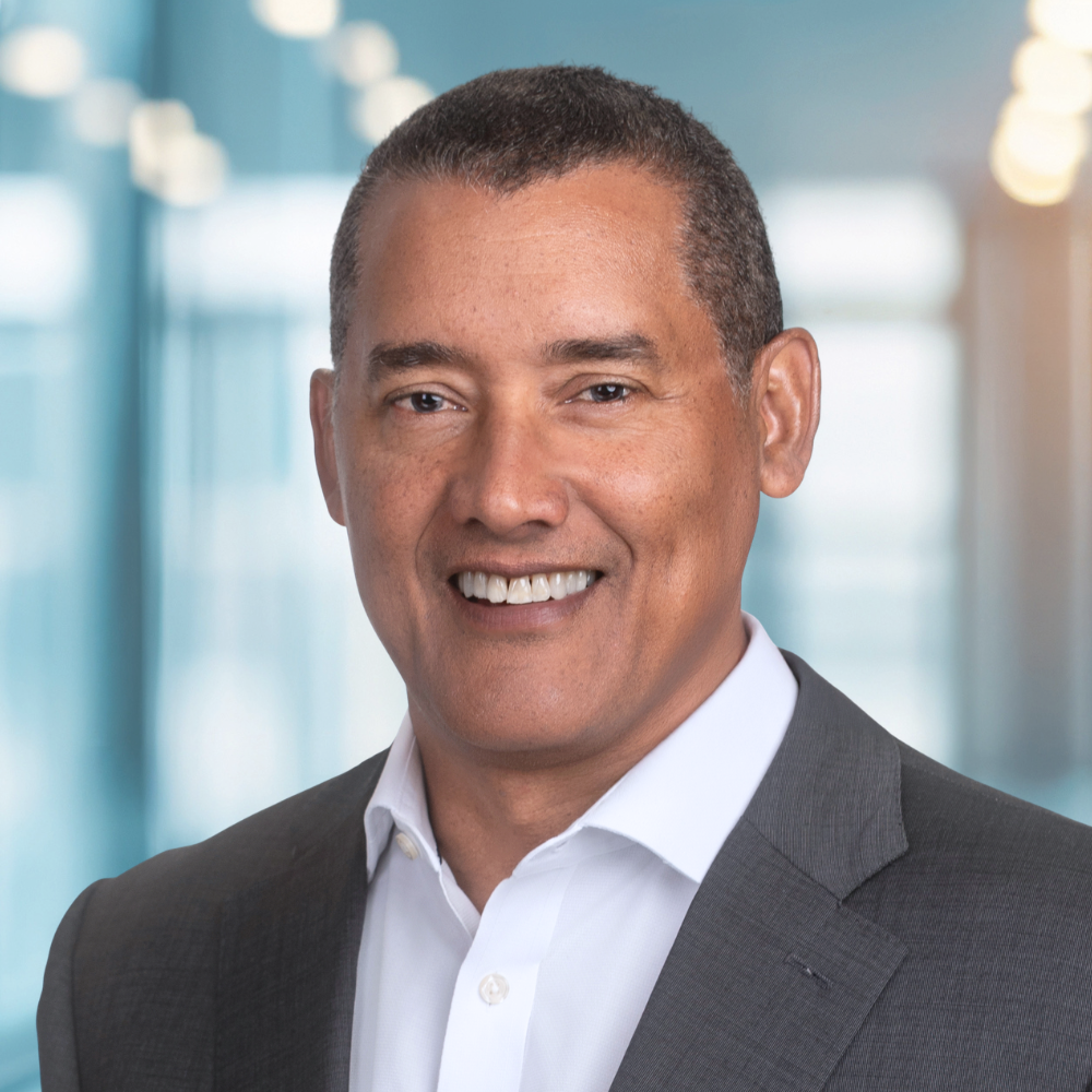 Professional headshot of a smiling man in a grey suit and white shirt against a blurred blue office background.