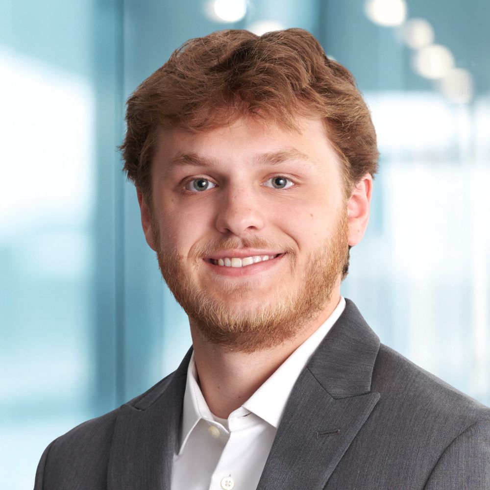 Head-and-shoulders portrait of a smiling man in a gray suit against a blue office background.