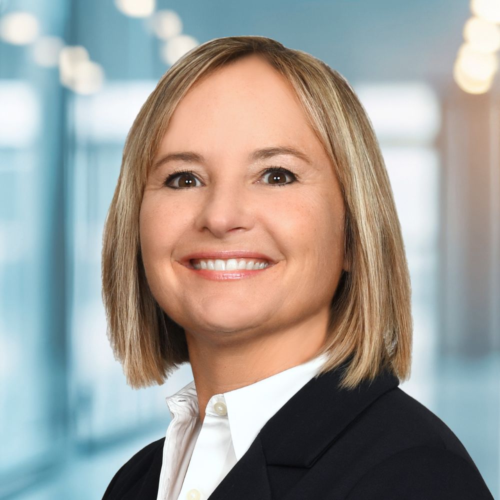 Professional headshot of a smiling woman in a black blazer and white shirt, neutral office background behind her.