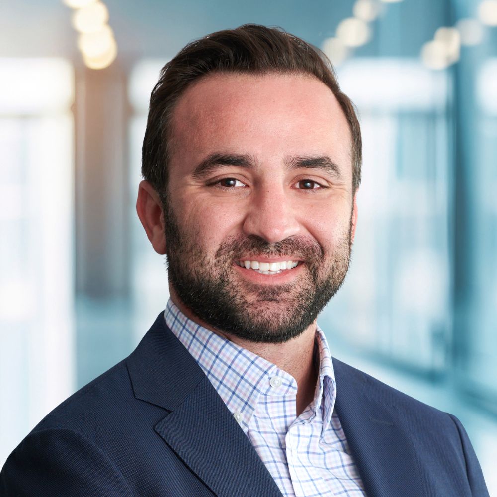 Professional headshot of a smiling man in a navy blazer and checkered shirt, in a bright office setting.