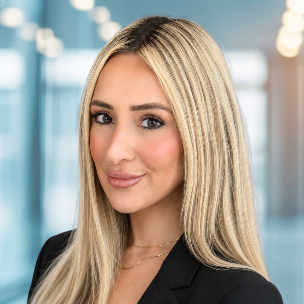 Professional headshot of a blonde woman in a black blazer, looking at the camera with a slight smile in a bright office setting.