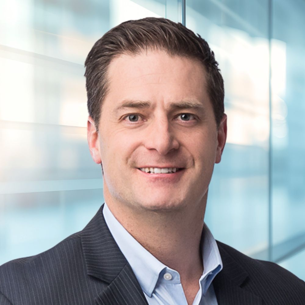 Professional headshot of a smiling man in a dark pinstripe suit against a glass office backdrop.
