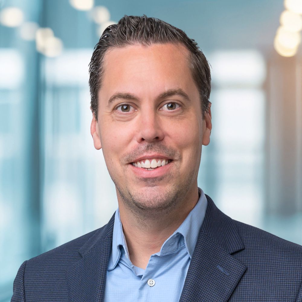 Smiling man in a blue shirt and dark blazer in a bright office hallway, professional headshot.