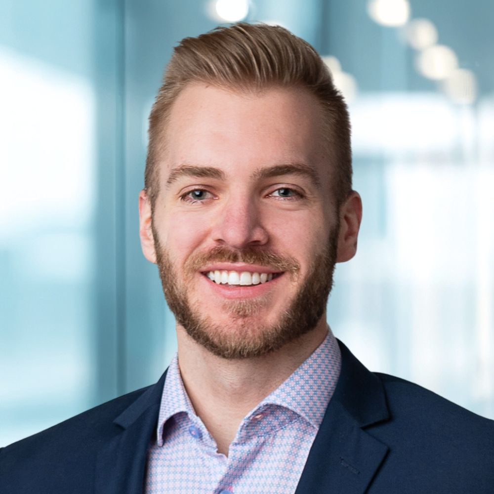 Head-and-shoulders portrait of a smiling man with blonde hair and a beard, wearing a navy blazer and light pink checkered shirt, in a bright office setting.