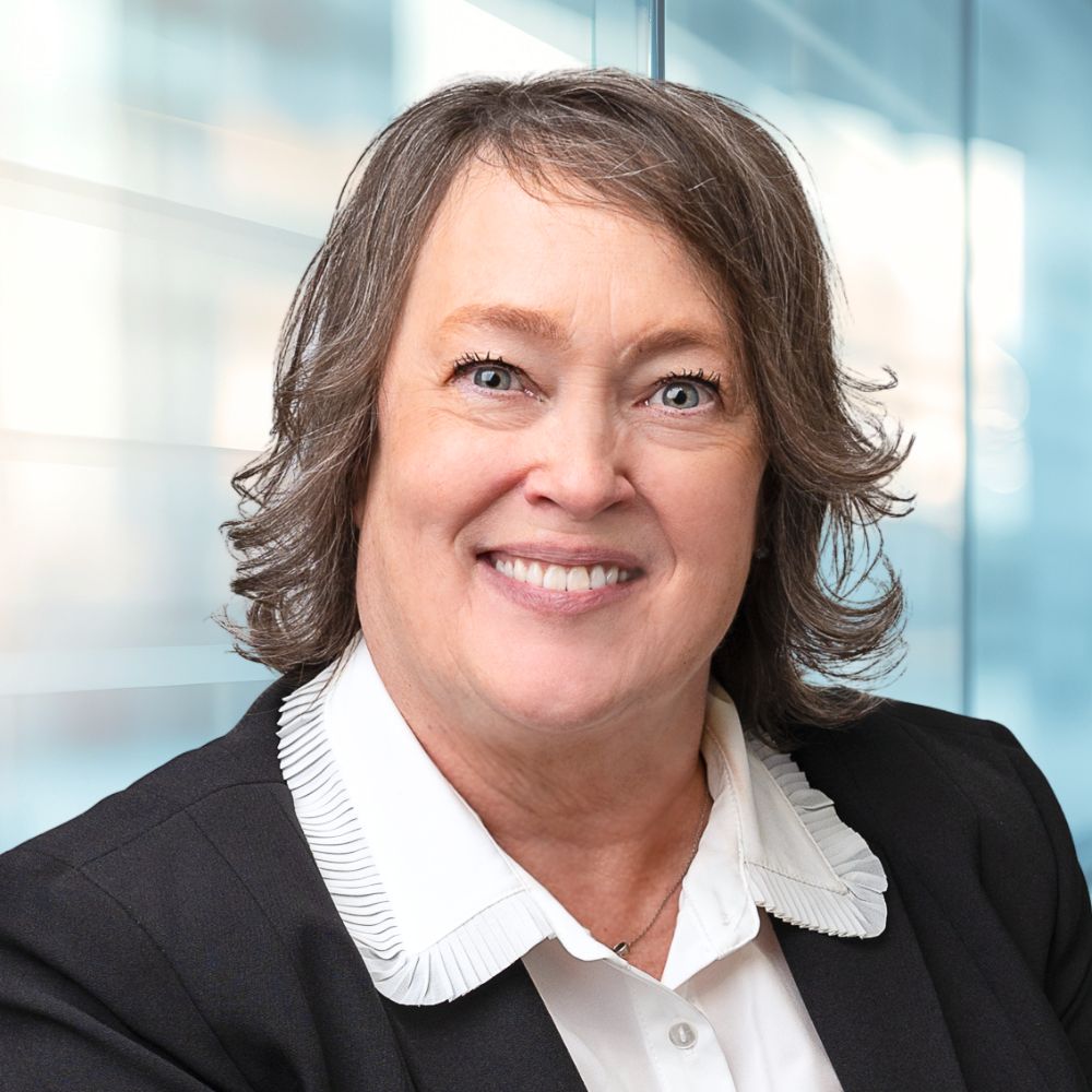 Smiling professional woman in a black blazer and white ruffled blouse, posing in a bright office with glass walls.