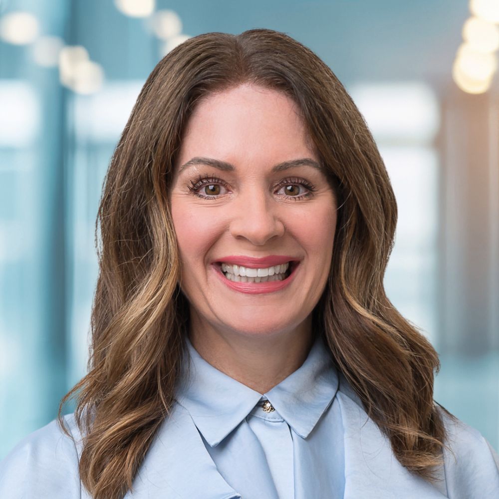 Smiling woman with brown wavy hair wearing a light blue blouse, in a modern office hallway.