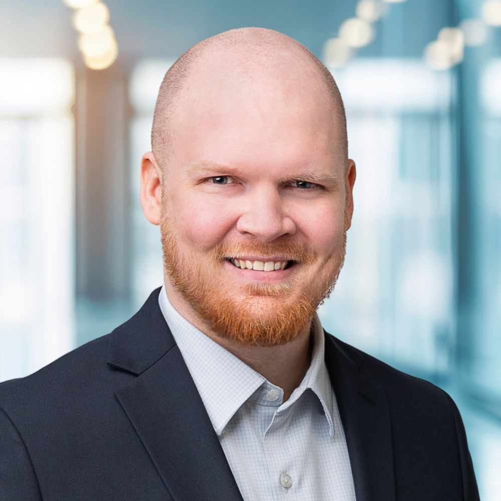 Professional headshot of a smiling bald man with a red beard in a dark blazer and light checkered shirt, office backdrop in soft focus