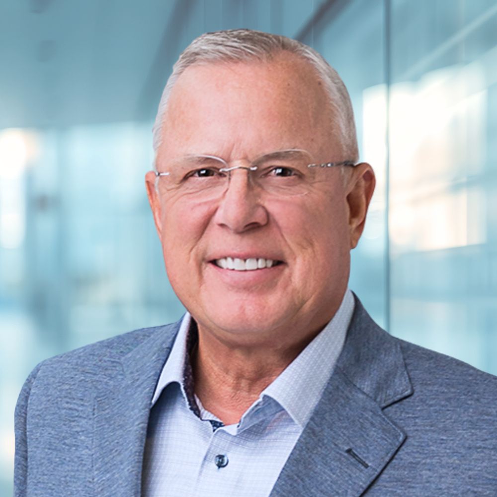 Portrait of a smiling older man wearing glasses, a light blue shirt, and a blue blazer in a glass-walled office hallway backdrop.