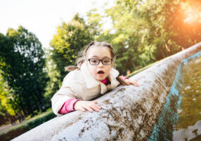 Portrait of beautiful young girl with two braids in eyeglasses, white coat looking aside smiling enjoying good sunny weather in autumn park outdoor. Sun glare effect