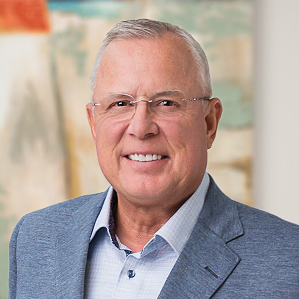 Smiling older man with gray hair and glasses, wearing a blue blazer and light dress shirt.