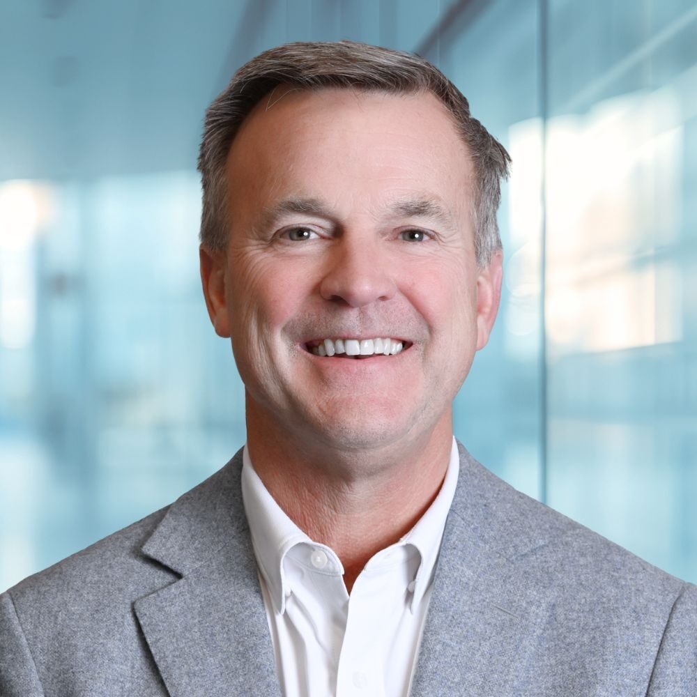 Professional headshot of a smiling man in a light gray blazer and white shirt, office backdrop in soft focus.