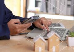 Person in a navy suit uses a smartphone while typing on a laptop at a desk, with small house models in the foreground.