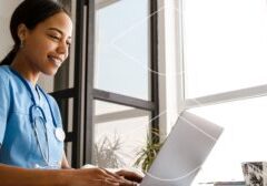 Nurse in blue scrubs with stethoscope typing on a laptop by a window in a bright clinic.