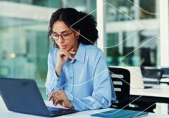 Professional woman wearing glasses, focused on a laptop at a bright office desk.